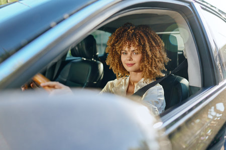 Smiling woman driving car with seatbelt fastened, curly hair and casual clothing, bright daylight and modern vehicle interior visible.の写真素材