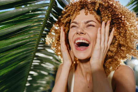 joyful woman with curly hair laughing outdoors near tropical leaves under bright sunlight, experiencing happiness and natural beauty in summer.の写真素材
