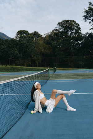 Young female tennis player enjoying her break on a blue court, wearing a sporty outfit and white sneakers, radiating joy and relaxation during sunny weatherの写真素材