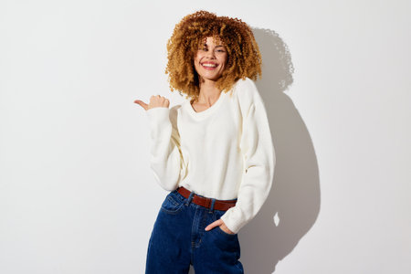 Cheerful young woman with curly hair in a cozy white sweater posing against a light background,...