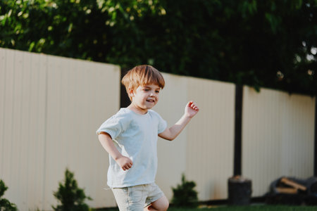 Happy boy running outdoors in a green garden with playful energy, enjoying a sunny day surrounded by nature and a cozy wooden fence.の写真素材