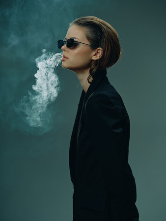 Stylish woman in black jacket wearing sunglasses exhales smoke against dark background with moody...