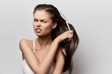 Frustrated woman with tangled hair, holding a comb against a light gray background, displaying a...