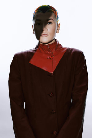 Woman with short hair wearing a dark red high-collar coat, dramatic lighting casting shadows on her...