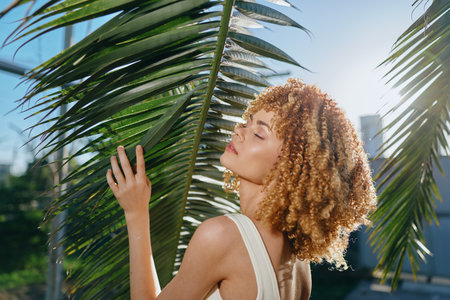 young woman with curly hair enjoying sunlight behind palm leaves, natural beauty, summer vibe, outdoor portrait with warm lightの写真素材