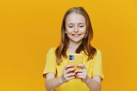 Happy girl with smartphone wearing yellow shirt against yellow background, smiling and enjoying technology, perfect for vibrant lifestyle and social media themesの写真素材