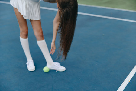 Young female tennis player in a white outfit reaching for a tennis ball on a blue court, showcasing athleticism and focusの写真素材
