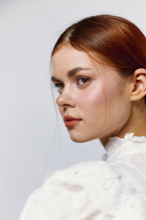 Elegant portrait of a young woman with red hair in a white vintage dress, conveying confidence and serenity on a soft, neutral backgroundの写真素材