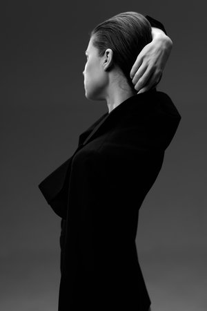 Black and white portrait of a woman with short hair wearing a dark coat, posed with her arm raised behind her head against a solid gray background, evoking elegance and poise.の写真素材