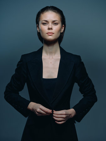 serious woman in black blazer adjusting jacket with focused expression against dark gray background in studio lighting, elegant and poised with natural makeupの写真素材