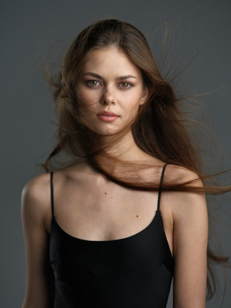 serious woman with long brown hair wearing black dress facing forward with wind blowing strands softly against gray studio background and calm expressionの写真素材