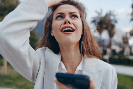 Businesswoman expressing frustration outdoors, wearing a light blazer and looking up with concern amidst a vibrant city backdrop with greenery.の写真素材