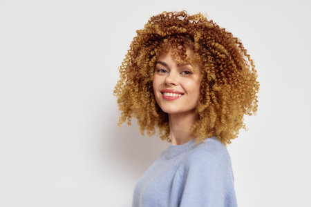 Beautiful woman with curly hair smiling against a light background showcasing her natural beauty and vibrant personality in a soft pastel colored sweaterの写真素材