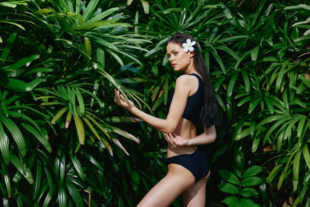 Exotic Asian Woman in Black Swimsuit Poses in Lush Tropical Jungle with Vibrant Green Plantsの写真素材