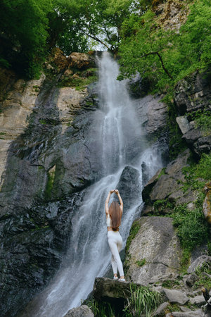 woman stretching near waterfall, natural outdoor scenery, green forest background, rocky cliff, peaceful atmosphere in summer nature settingの写真素材