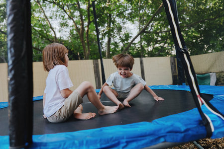 Two playful children enjoying a fun time on a trampoline, surrounded by lush greenery and bright sunlight. Capturing the joy of childhood and active play.の写真素材