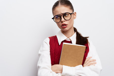 Student girl with glasses looking frustrated, holding a notebook against a plain white background, dressed in a red sweater and white shirt, conveying emotions of stress and concentrationの写真素材