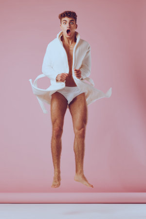 Excited young man jumping in white robe and underwear, expressing joy and surprise against a pink background. Fun and dynamic lifestyle concept.の写真素材