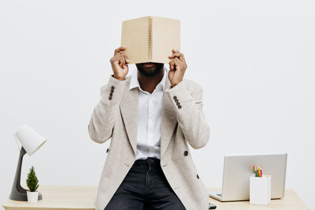 Professional male in casual blazer holding closed book in front of face, sitting at tidy modern...