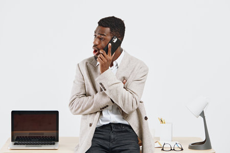 Businessman with headphones talking on mobile phone sits at desk with laptop and office supplies in bright modern workspace, concept of communication, technology, remote work, office environment, multitasking, professional attire.の写真素材