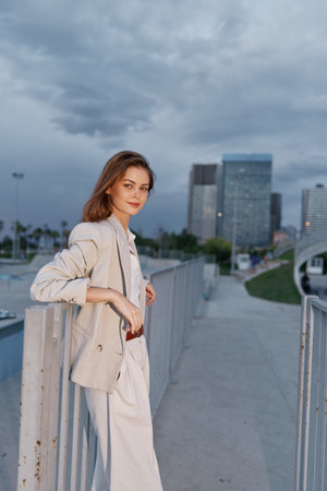 Young woman in beige business suit standing outdoors with skyscrapers background, confident professional woman in urban setting at dusk, business fashion conceptの写真素材