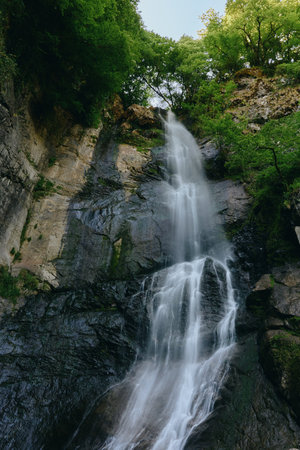 Stunning waterfall cascades down rocky cliffs surrounded by lush green forest and vibrant foliage,...