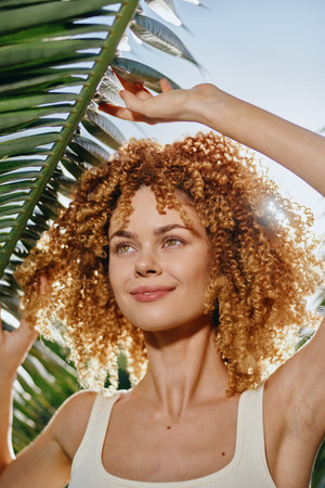 smiling woman with curly hair holds tropical palm leaf under bright sunlight, fresh summer vibe, natural beauty in casual white tank top outdoorsの写真素材