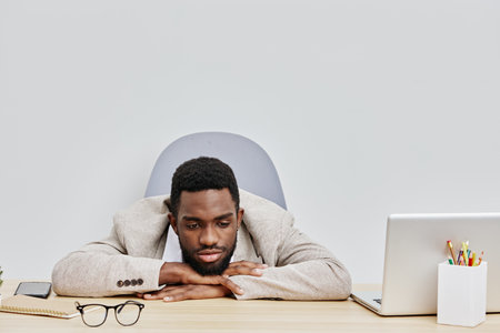 Focused young african-american man with beard resting head on folded arms at desk in office workspace, looking bored or tired, surrounded by laptop, glasses, and office supplies, professional workplace scene, modern style.の写真素材