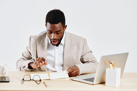 Professional young African American man wearing beige blazer and white shirt working at desk with...