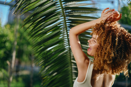 smiling woman with curly hair enjoying sunny tropical outdoors, wearing a white tank top, surrounded by green palm leaves, radiating happiness and warmth.の写真素材