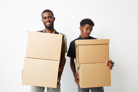 Two smiling African American men holding cardboard boxes in a bright, minimalistic setting. Their expressions show a mix of joy and playfulness, highlighting teamwork.の写真素材