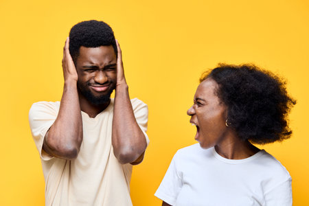 A young African couple expressing emotions, with the man covering his ears and the woman yelling, set against a vibrant yellow backgroundの写真素材