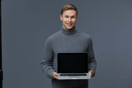 Young smiling caucasian man wear gray turtleneck sweater hold laptop computer present digital nomad concept on neutral gray background studio portraitの写真素材