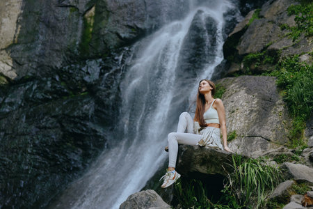 Woman sitting near waterfall in casual outfit relaxing outdoors, surrounded by rocks and greenery, enjoying nature and calm atmosphere on sunny day.の写真素材