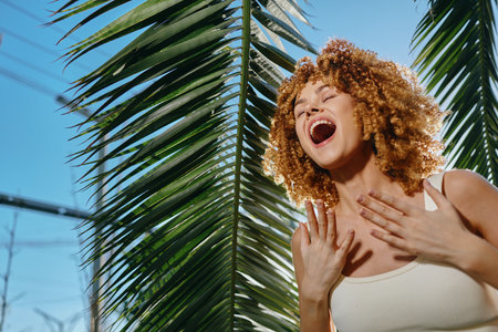 joyful woman with curly hair singing outdoors near tropical palm leaves under clear blue sky, wearing white tank top and expressing happiness.の写真素材