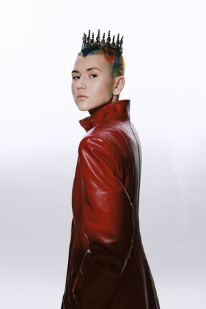 Woman with colorful hair and spiked crown wearing a red leather coat, posing confidently against a...