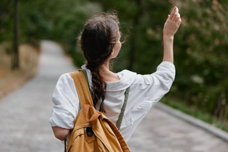 A young woman waving goodbye while walking on a tree lined path, showcasing casual, stylish outdoor attire and a sense of optimismの写真素材