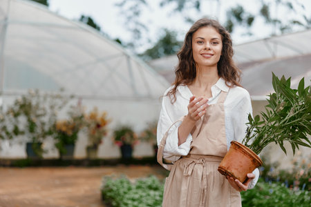Young woman with curly hair wearing a white shirt and apron, holding a potted plant in a greenhouse, embodying a love for gardening and natureの写真素材