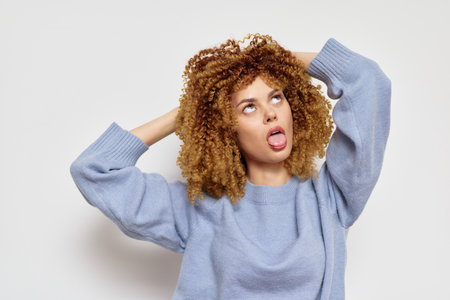 Young woman with curly hair showing playful expression, wearing a cozy blue sweater against a light gray background, embodying fun and carefree attitude in lifestyle photographyの写真素材