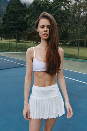 Young athletic woman in a white tennis outfit poses confidently on the court, showcasing a fit physique and determinationの写真素材