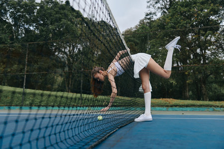 Young female tennis player adjusting the net on a beautiful outdoor court, showcasing athleticism and determination in a stylish white outfit, capturing the essence of sports and competitionの写真素材