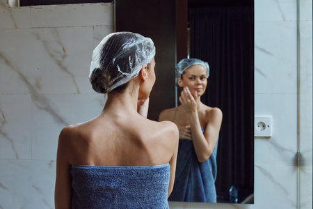 Woman with shower cap wrapped in blue towel touching face in bathroom mirror, skincare routine, personal care, wellness moment, morning self-care.の写真素材
