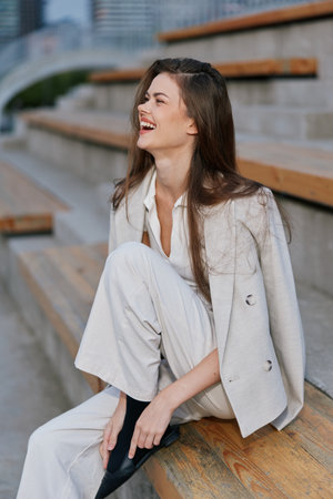Woman laughing outdoors in stylish business outfit sitting on wooden steps with urban background, showing joyful emotion and casual confidenceの写真素材