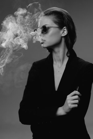 Black and white portrait of woman wearing sunglasses exhaling vapor smoke holding vape device with stylish attitude in studio lighting and dark backgroundの写真素材