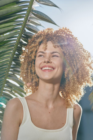 smiling woman with curly hair enjoys sunny tropical background, warm natural light enhancing bright joyful mood and relaxed summer atmosphere outdoorsの写真素材