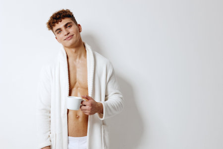 Smiling young man in a white bathrobe, holding a cup of coffee, stands against a light background. Morning relaxation and self-care concept.の写真素材