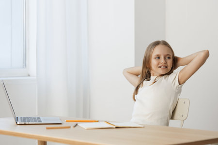 Cheerful young girl relaxing at desk with laptop, enjoying study time, minimalistic background, bright white tones, casual outfit reflecting confidenceの写真素材