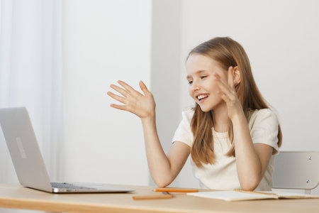 Happy young girl waving at her laptop during an online call, wearing a casual white shirt, in a...
