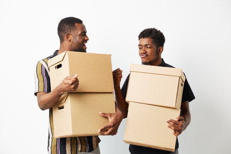 Two smiling African American men share a moment while carrying cardboard boxes, displaying teamwork and joy in a minimalist setting. Concept: friendship and collaboration.の写真素材