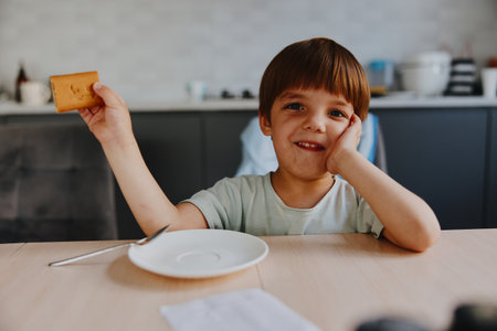 Happy boy enjoying a snack, looking excited with a cookie in hand and a joyful expression, creating a cheerful moment in a cozy kitchen setting.の写真素材